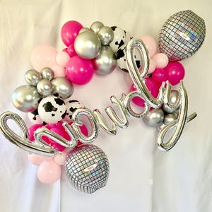 K&ouml;nnte beinhalten: Ein silberner Ballonbogen, der "Howdy" buchstabiert, mit rosa, silbernen und schwarz-wei&szlig;en Kuhmuster-Ballons.