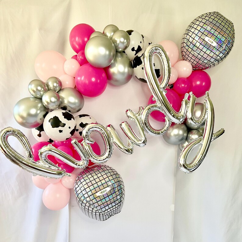 K&ouml;nnte beinhalten: Ein silberner Ballonbogen, der "Howdy" buchstabiert, mit rosa, silbernen und schwarz-wei&szlig;en Kuhmuster-Ballons.