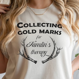 May include: A white t-shirt with black text that reads "COLLECTING GOLD MARKS for Jamlin's therapy" and a black graphic of antlers.