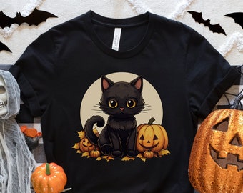 Könnte beinhalten: Schwarzes T-Shirt mit einer niedlichen schwarzen Katze, die einen Kürbis-Hut trägt und von Kürbissen und Blättern umgeben ist. Die Katze sitzt vor einem Vollmond.