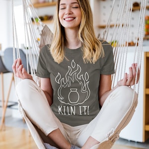 Puede incluir: Una camiseta gris con un gráfico blanco de un horno en llamas y el texto "KILN IT!". La camiseta la lleva una persona sentada en una silla colgante. La persona sonríe con los ojos cerrados.