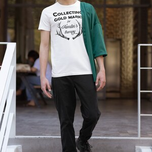 May include: A person wearing a white t-shirt with the text "COLLECTING GOLD MARKS for Tamlin's therapy" and a green jacket. The person is standing on a set of stairs.