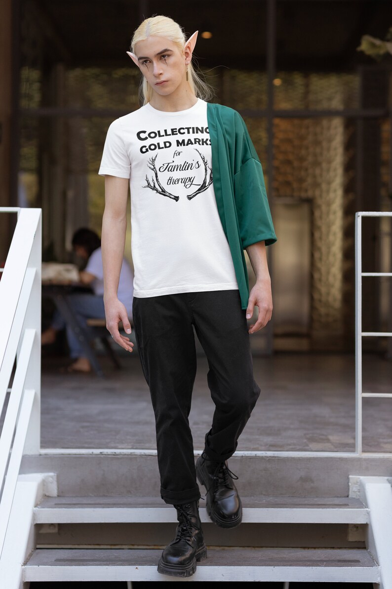 May include: A person wearing a white t-shirt with the text "COLLECTING GOLD MARKS for Tamlin's therapy" and a green jacket. The person is standing on a set of stairs.