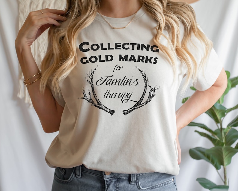 May include: A white t-shirt with black text that reads "COLLECTING GOLD MARKS for Jamlin's therapy" and a black graphic of antlers.