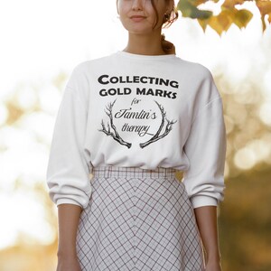 May include: White sweatshirt with black text that reads "COLLECTING GOLD MARKS for Jamlin's therapy" with a black antler graphic.