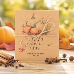 May include: A square tan card tied with twine featuring a watercolor illustration of pumpkins and autumn leaves. The text reads: Love is the spice of life, Shana & Cameron, 10.12.24. The card is decorated with small red leaf accents.