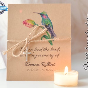 May include: A brown card with a hummingbird and a tulip illustration. The card has a silver cross charm attached with twine and the text "Please feed the birds in loving memory of Donna Rollins 2/1/28 - 6/11/19" printed on it. The card is next to a white tea light candle.