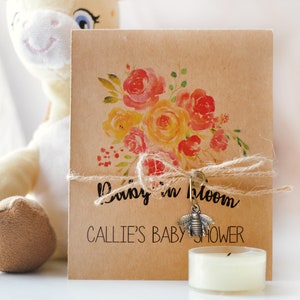May include: A kraft paper baby shower invitation with a watercolor floral design in shades of red, orange, and yellow. The invitation is tied with twine and features the text "Baby in bloom" and "Callie's Baby Shower". A small bee charm hangs from the twine.