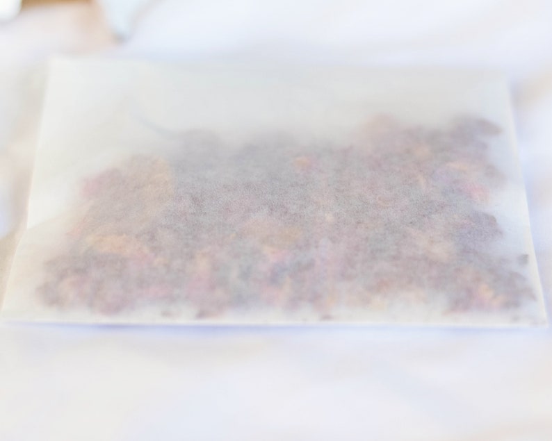 Op de afbeelding: Een rechthoekige, doorschijnende papieren verpakking gevuld met gedroogde, kleurrijke bloemblaadjes. De bloemblaadjes zijn een mix van roze, rode en oranje tinten, zichtbaar door het delicate papier. De verpakking ligt op een wit oppervlak.