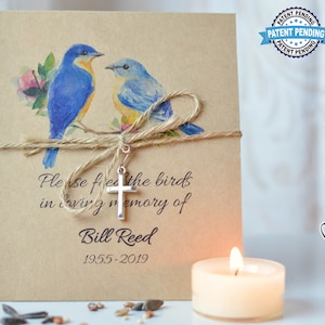 May include: A brown paper card with a watercolor illustration of two bluebirds perched on a branch. The card has the text "Please feed the birds in loving memory of Bill Reed 1955-2019". A silver cross charm hangs from a twine bow tied around the card. The card is next to a white candle.