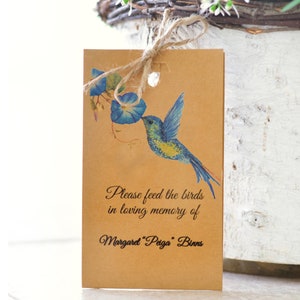 Op de afbeelding: Een bruine kaart met een aquarelkolibrie en blauwe bloemen. De tekst luidt: "Please feed the birds in loving memory of Margaret 'Peiga' Binns." De kaart is vastgebonden met touw en de woorden "FEED THE BIRDS FUNERAL FAVOR" zijn verticaal afgedrukt.