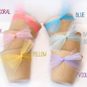 May include: A collection of small, beige pots adorned with colorful tulle bows. The bows are in shades of coral, pink, yellow, blue, baby blue, and violet. The word labels for each color are visible.