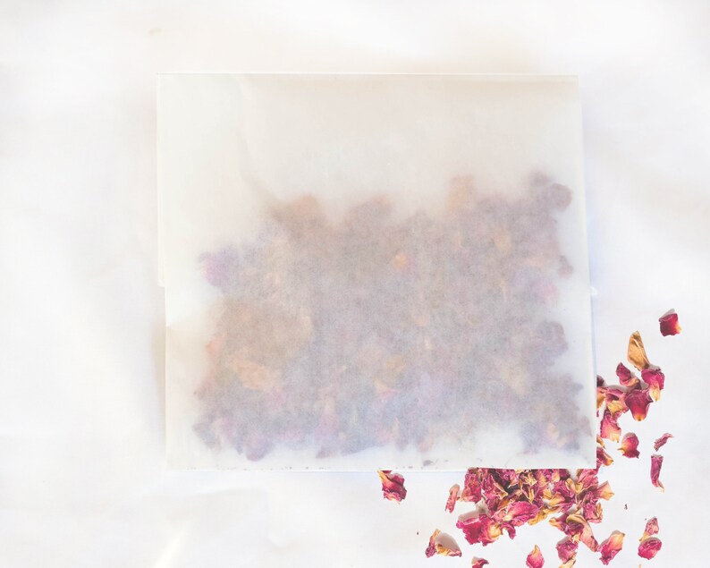 Op de afbeelding: Een vierkant, doorschijnend papieren pakket gevuld met gedroogde roze rozenblaadjes, met wat bloemblaadjes eromheen verspreid. Het pakket staat op een witte achtergrond, wat een strakke en eenvoudige uitstraling cre&euml;ert.