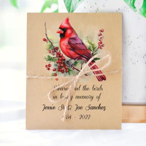 May include: A rectangular kraft paper card featuring a vibrant red cardinal perched on a branch with red berries and green leaves. The text reads: Please feed the birds in loving memory of Jessie Sr & Joe Sanchez 1954 - 2022. A jute twine bow is tied around the card.