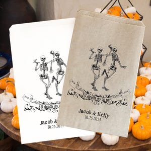 Op de afbeelding: Twee skeletten die dansen met de tekst "Til death do us part" en "Jacob & Kelly 10.25.2025" op een bruine papieren zak. De zak is versierd met een bloemmotief.