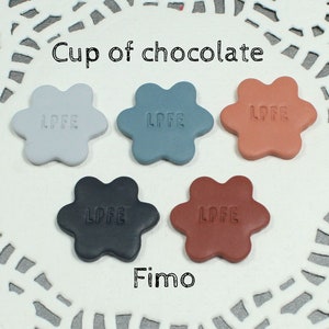 Może przedstawiać: Pięć kwiatów z polimerowej gliny w różnych kolorach: jasnoszary, niebieski, pomarańczowy, czarny i brązowy. Każdy kwiat ma wytłoczone słowo "LPFE". Kwiaty są ułożone na białej koronce. Tekst "Cup of chocolate" i "Fimo" jest napisany nad i pod kwiatami.