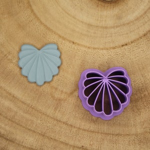 May include: Two heart-shaped cookie cutters. One is light blue and the other is purple. Both cutters have a scalloped edge and a fan-like design in the center. The cutters are made of plastic and are on a wooden surface.