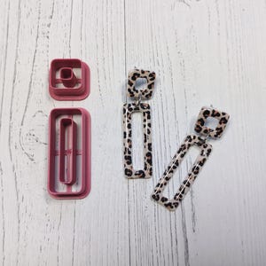 Puede incluir: Dos pares de pendientes con diseño de estampado de leopardo. Los pendientes están hechos de un material blanco con manchas negras. Los pendientes tienen la forma de un rectángulo con un cuadrado más pequeño en la parte superior.