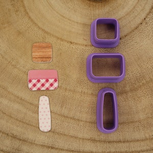 May include: Set of four purple plastic cookie cutters on a wooden surface. The cutters are shaped like a square, rectangle, oval, and a long rectangle. There are also three small decorative pieces with pink and white checkered patterns.