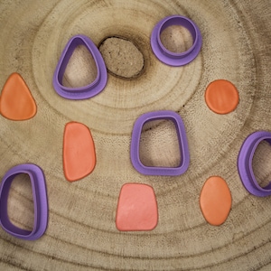 Könnte beinhalten: Ein Satz von acht violetten und orangefarbenen Ausstechern auf einer Holzoberfläche. Die Ausstecher sind in Form von Kreisen, Ovalen und einer Träne.