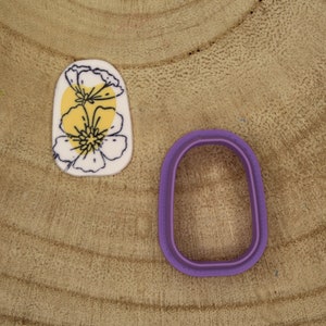 Peut inclure: Un emporte-pièce ovale en plastique violet. Un biscuit blanc avec un motif floral jaune et noir est en arrière-plan.