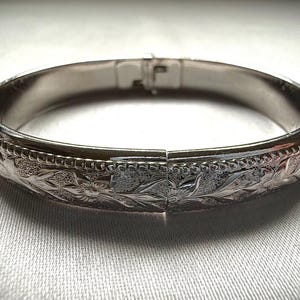 May include: A silver bangle bracelet with a floral design. The bracelet is hinged and has a smooth, polished finish.