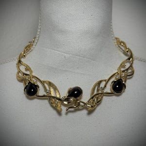 May include: A gold-tone necklace with a delicate, leafy design and three black gemstone accents. The necklace is on a white background.