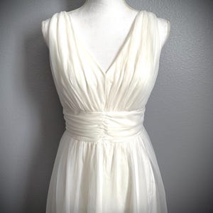 May include: Ivory-colored sleeveless dress with a V-neckline and a gathered waist. The dress is displayed on a mannequin against a gray background. The fabric appears to be lightweight and flowing, with a subtle sheen.