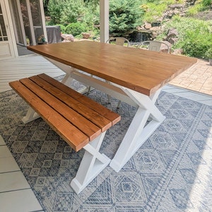 May include: A white wooden table with a brown wooden top and a matching bench. The table and bench are on a blue and white patterned rug. The table and bench are on a porch with a view of a backyard.