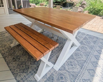 Puede incluir: Una mesa de madera blanca con una tapa de madera marrón y un banco a juego. La mesa y el banco están sobre una alfombra de estampado azul y blanco. La mesa y el banco están en un porche con vistas a un jardín.