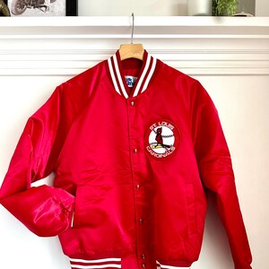Op de afbeelding: Rood satijnen honkbaljack met witte en rode strepen aan de zoom. Het jack heeft een knoopsluiting en een St. Louis Cardinals logo op de linkerborst.
