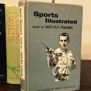 Könnte beinhalten: Ein blaues Vintage-Buch mit dem Titel "Sports Illustrated Book of Wet-Fly Fishing" mit einer Schwarzweißillustration eines Mannes beim Fliegenfischen. Das Buch wurde von den Redakteuren von Sports Illustrated herausgegeben.