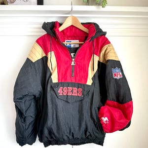 Puede incluir: Chaqueta vintage de los 49ers en negro, rojo y dorado con capucha y bolsillo delantero. La chaqueta presenta el logotipo de la NFL en la manga y el nombre del equipo bordado en la parte delantera. La chaqueta está colgada en una percha de madera.