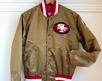 Può includere: Un giubbotto bomber in raso dorato con colletto, polsini e fascia in vita a righe rosse e bianche. La parte anteriore presenta una toppa con il logo dei San Francisco 49ers sul petto a sinistra, un piccolo stemma NFL sulla manica e bottoni.