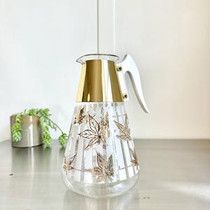May include: A clear glass pitcher with a gold top and a white handle. The pitcher has a white and gold leaf design.