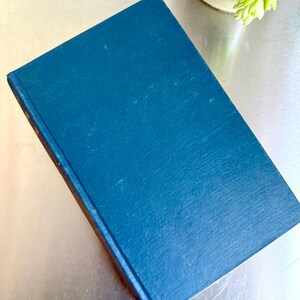 May include: A hardback book with a textured, dark teal cover. The book is angled, showing a glimpse of the spine. A small succulent plant in a beige pot is visible in the background.