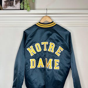 Puede incluir: Una chaqueta de béisbol de satén azul oscuro con rayas amarillas y blancas en el cuello y los puños. La parte posterior de la chaqueta presenta un gran logotipo bordado "NOTRE DAME" en amarillo y blanco.