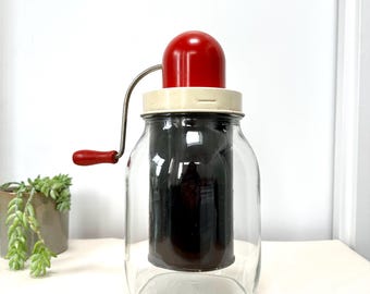 Puede incluir: Un molinillo de alimentos manual vintage rojo y blanco con un tarro de vidrio. El molinillo tiene una manivela y una tapa roja.