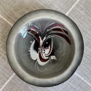 May include: A round, gray ceramic bowl featuring a hand-painted abstract design in white, black, and deep red. The center motif includes delicate, sweeping lines and small dots, with a signature visible near the bottom edge of the interior surface.