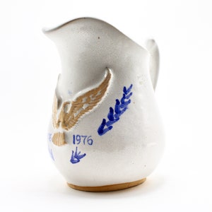 May include: White ceramic jug with a handle and spout. The jug features a tan eagle design with blue accents and the year "1976" in blue. The jug has a speckled texture and a brown base.