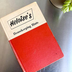 May include: Vintage book titled "Heloise's Housekeeping Hints." The book has a red and cream-colored cover. The title is in black lettering with a decorative logo. The book is slightly worn, suggesting it is an antique item. The book is angled on a metallic surface.