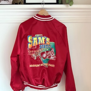 May include: Red satin bomber jacket with white and red striped trim. The back features a colourful graphic with the text "SAM'S STATION", a train, and the words "The Bad Boys of Central" and "Breakin' All The Rules".