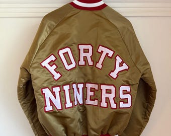 Può includere: Un giubbotto bomber in raso dorato con la scritta FORTY NINERS in lettere bianche con bordi rossi sul retro. Il giubbotto presenta colletto, polsini e orlo a coste con strisce rosse e bianche. È appeso a una gruccia di legno.