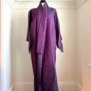 Pode incluir: Um quimono longo de cor roxo profundo com um padrão jacquard tonal de motivos circulares e linhas onduladas. A peça possui mangas largas e estruturadas, uma gola alta e um painel frontal que cai reto, exibindo um forro violeta vibrante.