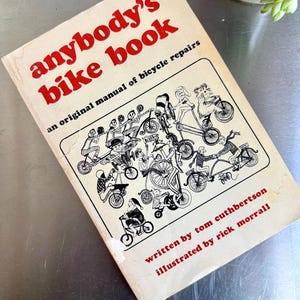 May include: Vintage "Anybody's Bike Book," an original manual of bicycle repairs. The cover features red text and illustrations of various bicycles and riders. Written by Tom Cuthbertson and illustrated by Rick Morrall.
