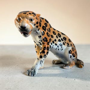 May include: A ceramic figurine of a leopard in a crouching position. The leopard has a tan coat with black rosettes and spots, a white underbelly, and a slightly open mouth showing teeth. The figure is finished with a glossy glaze.