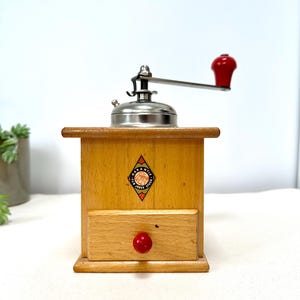 May include: A vintage wooden coffee grinder with a red handle and a drawer. The grinder has a silver metal top and a diamond-shaped label that reads "Garantie Jules Stodel".