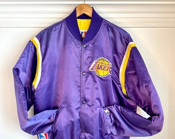 May include: Purple satin jacket with yellow and white accents. Features the Los Angeles Lakers logo on the chest and NBA logo on the sleeve. The jacket has a button-up front, two pockets, and a ribbed collar and cuffs.