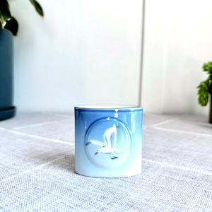Puede incluir: Una taza pequeña de cerámica azul y blanca con un diseño en relieve de un pájaro blanco en vuelo. La taza está sobre una superficie gris claro.