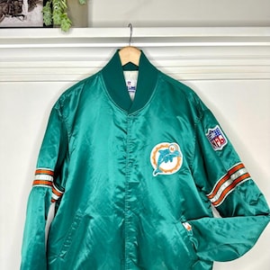 Puede incluir: Chaqueta de satén verde azulado con el logotipo de los Miami Dolphins y un parche de la NFL. La chaqueta tiene rayas naranjas y blancas en las mangas y cuello y puños verdes. Una prenda deportiva vintage.